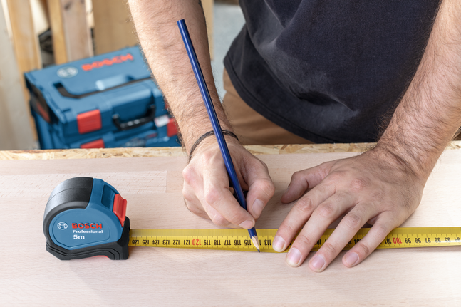 Oseba označuje mere na lesu z merilnim trakom in svinčnikom.